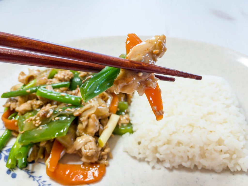 Chopsticks holding chicken and vegetable stir-fry over a plate of chicken stir-fry and white rice.