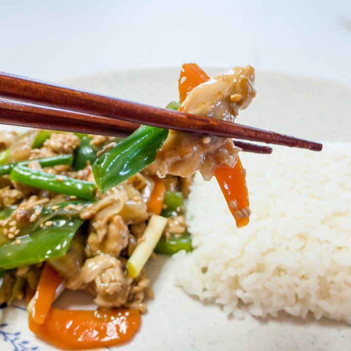 Chopsticks holding chicken and vegetable stir-fry over a plate of chicken stir-fry and white rice.