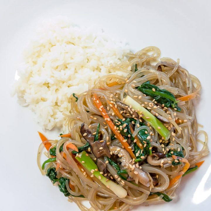 A closeup of ready to eat japchae (or chapchae) made with sweet potato starch noodles and vegetables that is next to a bit of white rice on a white plate.