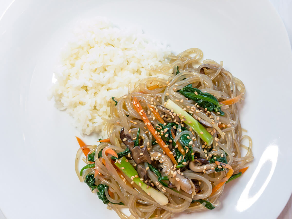 A closeup of ready to eat japchae (or chapchae) made with sweet potato starch noodles and vegetables that is next to a bit of white rice on a white plate. 