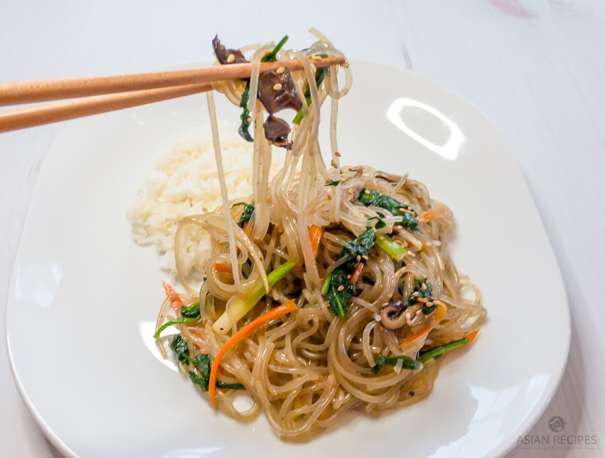 A closeup of ready to eat japchae (or chapchae) made with sweet potato starch noodles and vegetables that is next to a bit of white rice on a white plate. 